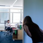 Junger Anwalt mit Kopfhörern am Schreibtisch in hellem Büro in der Kanzlei, während eine Frau im Vordergrund vorbeigeht